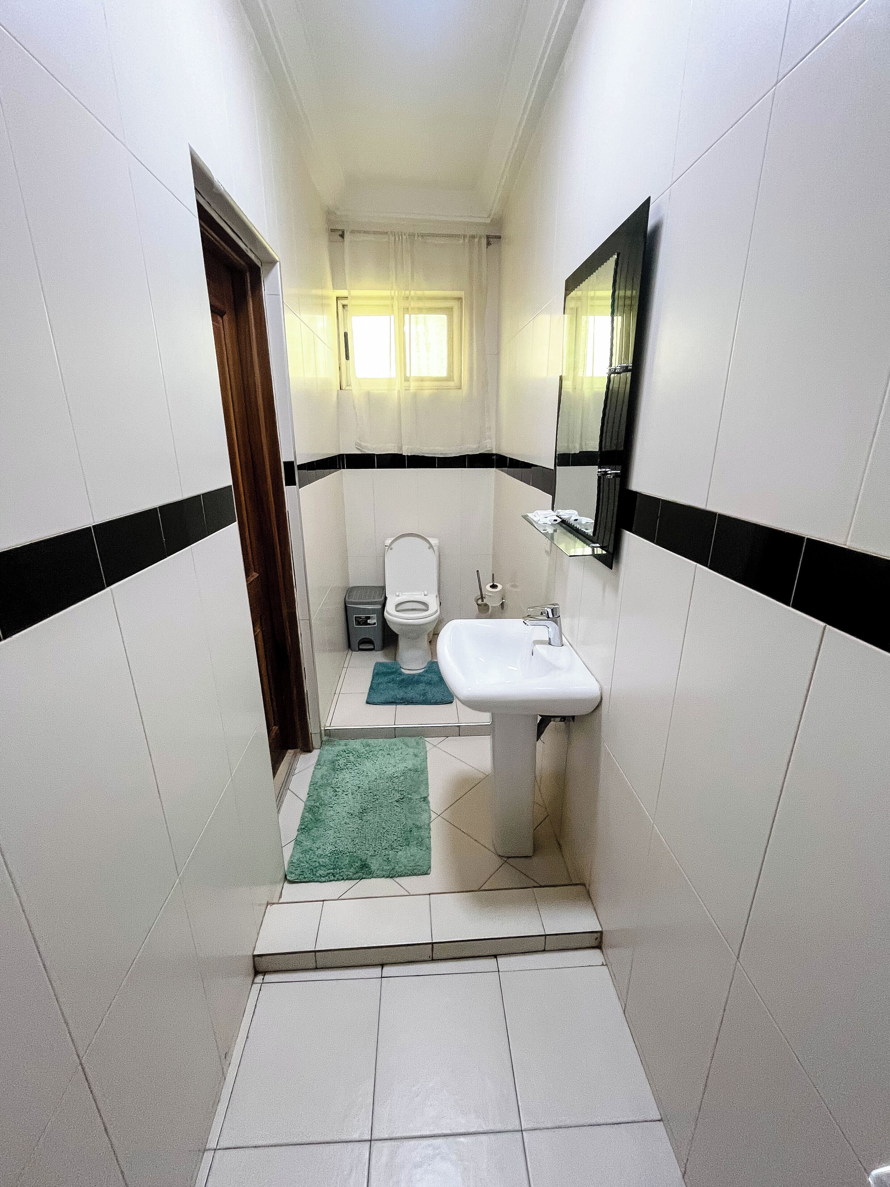 Un baño estrecho con azulejos blancos, un lavamanos blanco, un inodoro y un espejo. Hay dos alfombras en el suelo.