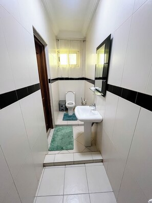 Un baño estrecho con azulejos blancos, un lavamanos blanco, un inodoro y un espejo. Hay dos alfombras en el suelo.