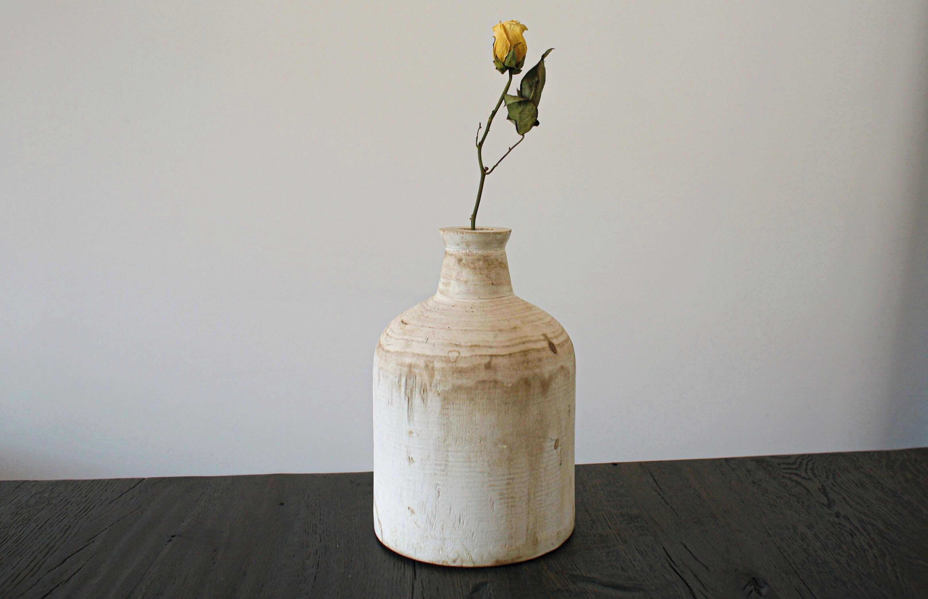 A white vase with a single yellow rose on a dark wooden table against a plain wall.
