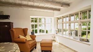 Ein Zimmer mit einem großen Fenster, einem braunen Ledersessel, einem kleinen Hocker und einem Tisch mit weißer Spitzenleinentuch.