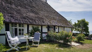 Ein traditionelles Haus mit einem Strohdach, einem Garten, zwei weißen Schaukelstühlen und einem Tisch mit einem Glas Wasser.