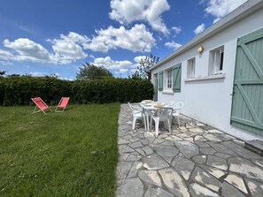 Une terrasse avec un sol en pierre et des chaises blanches, une petite table avec des assiettes, et un arbuste en arrière-plan.