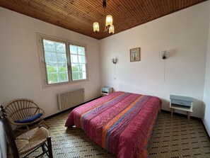 Une chambre avec un plafond en bois, un lit recouvert d’une courtepointe à motifs, une chaise en osier, une petite table et une fenêtre donnant sur un paysage verdoyant.
