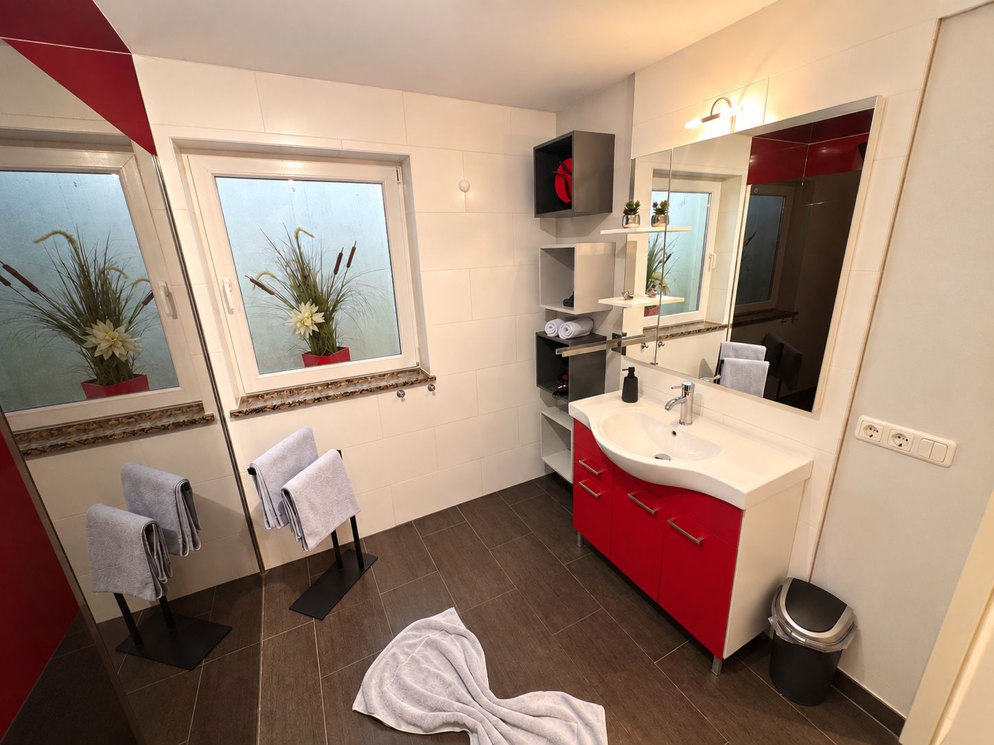 A modern bathroom with a large mirror, a white sink, red cabinets, and two potted plants on the windowsill.