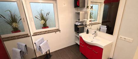 A modern bathroom with a large mirror, a white sink, red cabinets, and two potted plants on the windowsill.