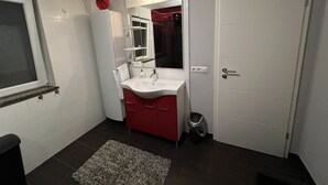 Un baño con lavamanos blanco, mueble rojo y espejo. Se ve una ventana con marco blanco y una puerta con manija.