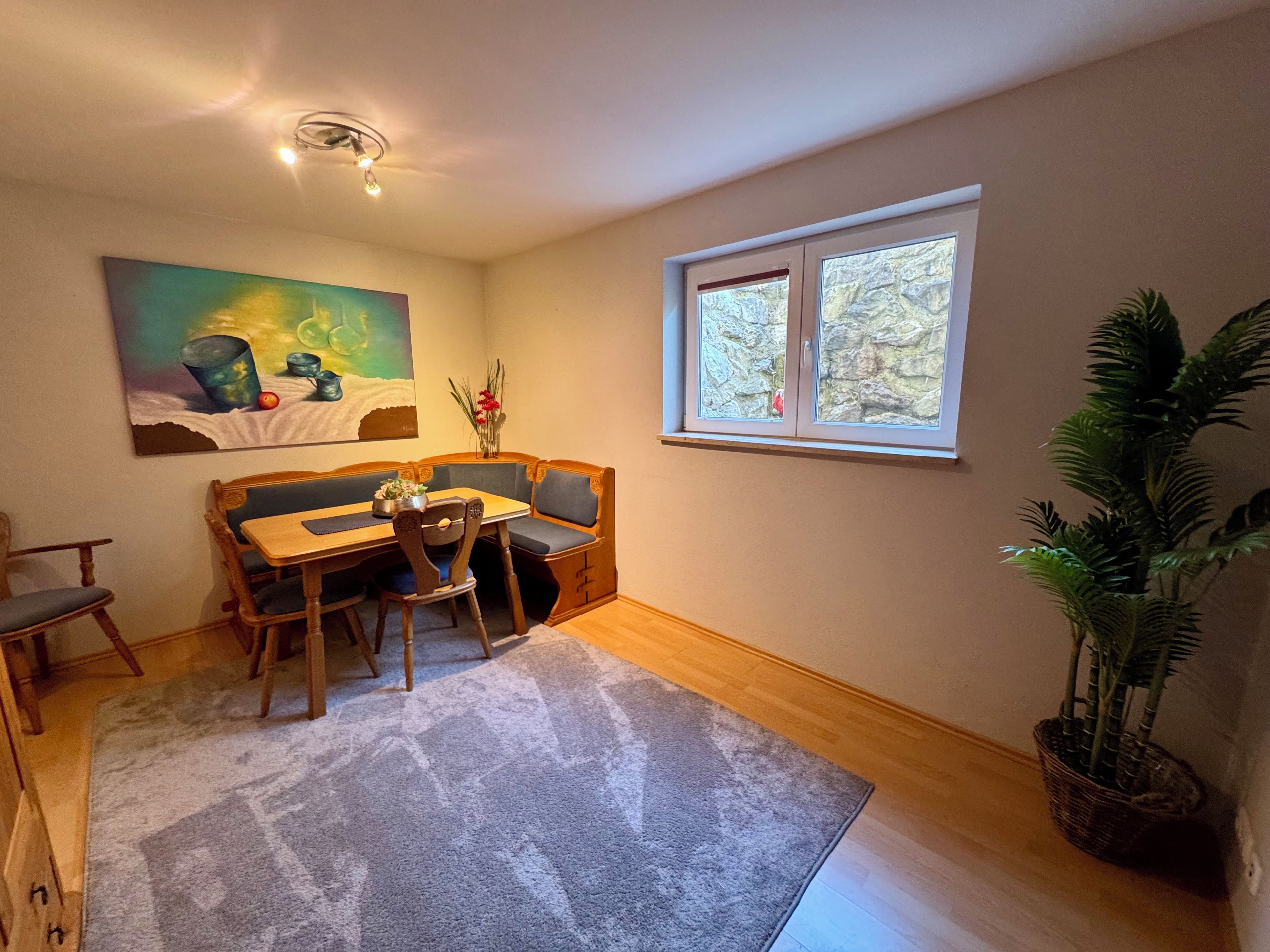 A dining area with a wooden table and chairs, a painting on the wall, a window with a view, and a potted plant.