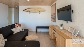 Un salon avec un canapé foncé, un banc en bois, une planche de surf accrochée au mur, un téléviseur et une console en bois sur laquelle est posée une plante.