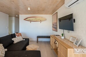 A living room with a dark sofa, a wooden bench, a surfboard on the wall, a TV, and a wooden console with a plant.