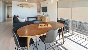 Un salon moderne avec une table à manger en bois, des chaises noires, un canapé, une planche de surf accrochée au mur, un téléviseur et un balcon avec vue.