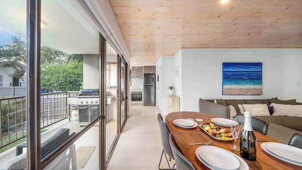 Une cuisine moderne donnant sur le jardin, un coin repas avec une table dressée pour deux personnes, et un salon avec un canapé et un tableau représentant l’océan.