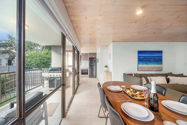 A modern kitchen with a view to the backyard, a dining area with a table set for two, and a living area with a couch and a painting of the ocean.