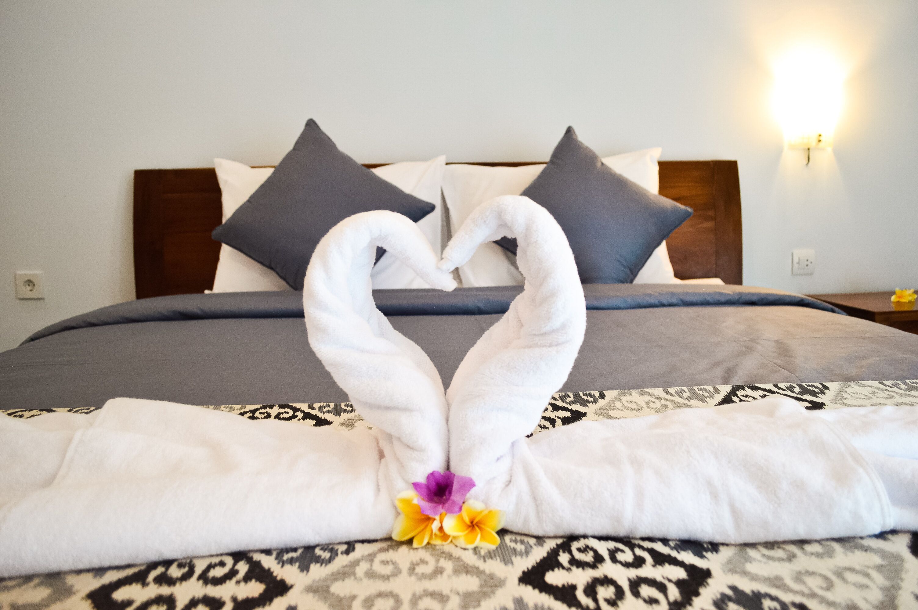A bed with a towel arranged in a heart shape, a patterned bedspread, and a wall-mounted light fixture.
