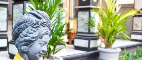 A statue of a woman holding a book, set in a garden with potted plants and a building with columns in the background.