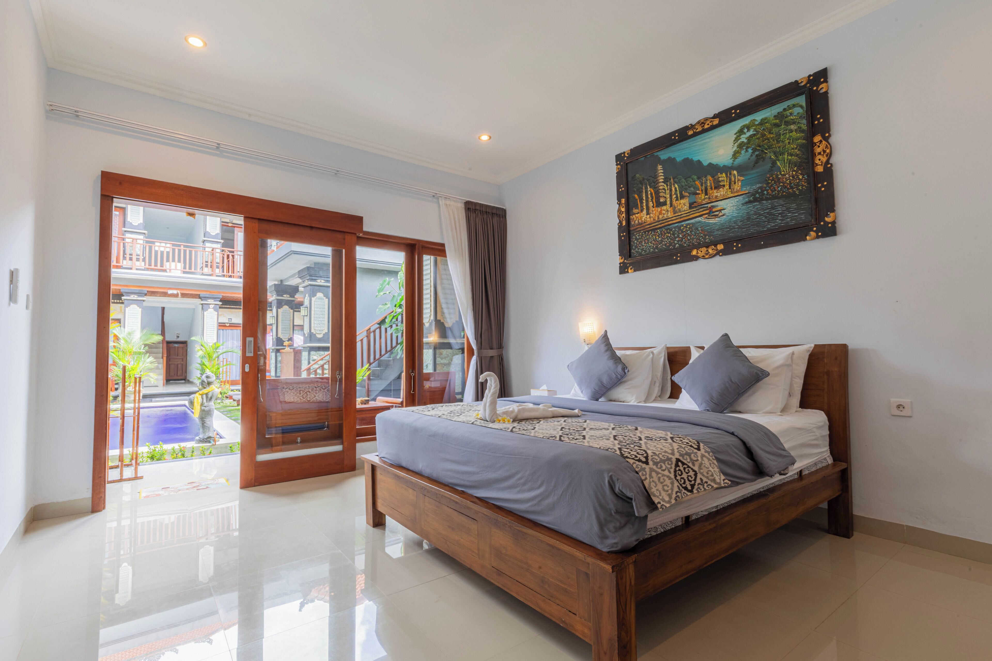 A bedroom with a wooden bed, a painting on the wall, and a view of a balcony with plants.