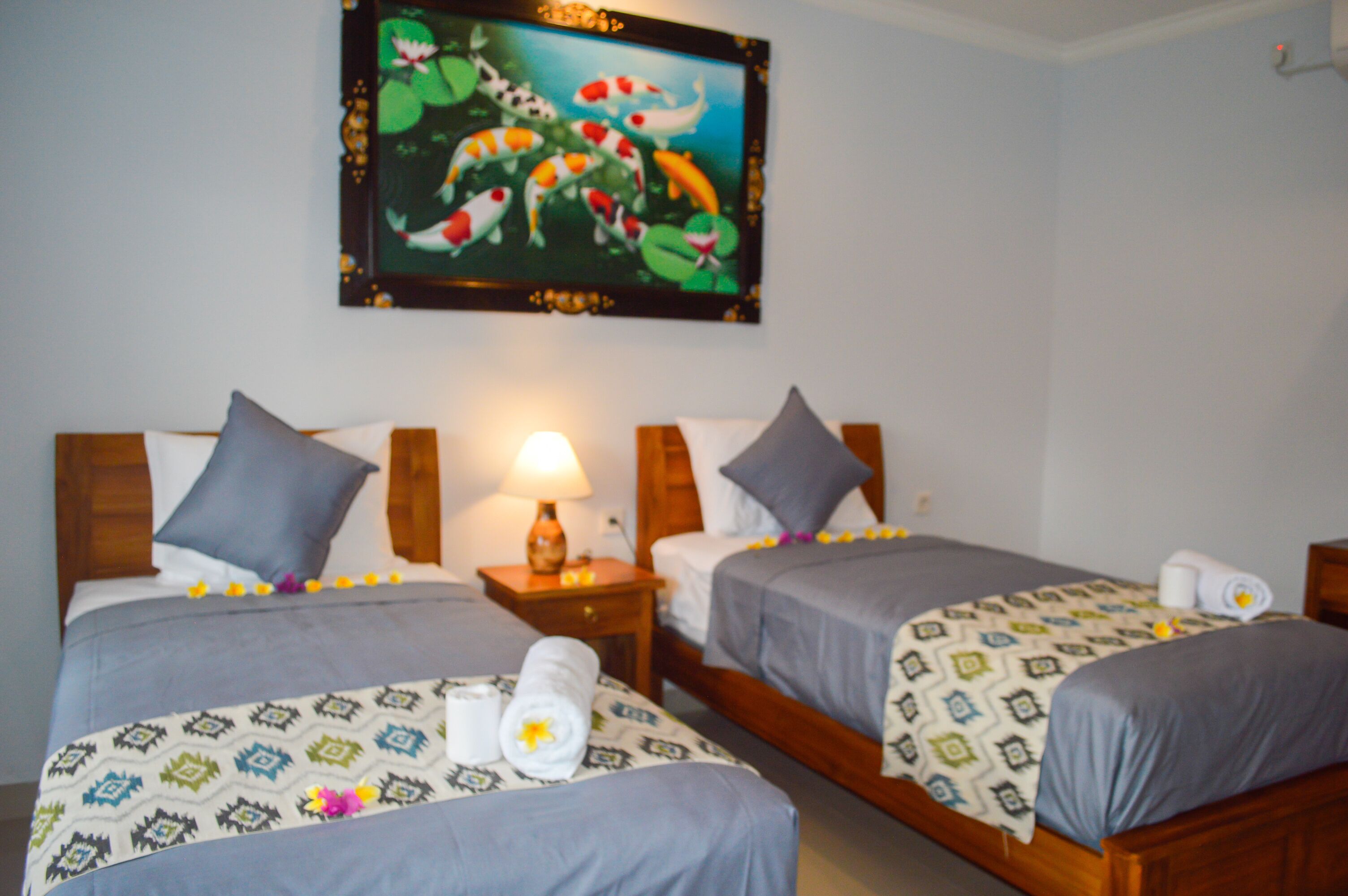 A hotel room with two beds, each with a patterned bedspread and pillows. There is a painting on the wall above the beds, a bedside table with a lamp, and a small nightstand.