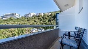 A balcony with a table and chairs, a view of a hillside with buildings, and parked cars.