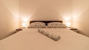 Ein ordentlich bezogenes Bett mit gemusterten Kissen und einer zusammengerollten Decke, zwei Nachttischlampen und ein hölzernes Kopfteil in einem Raum mit schlichten Wänden.