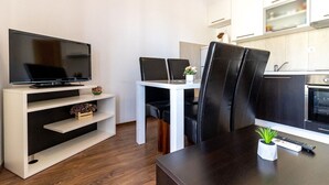 Ein modernes Wohnzimmer mit einem Flachbildfernseher auf einem weißen Schrank, einem Esstisch mit zwei Stühlen sowie einer Küchenzeile mit Schränken und einem Herd.