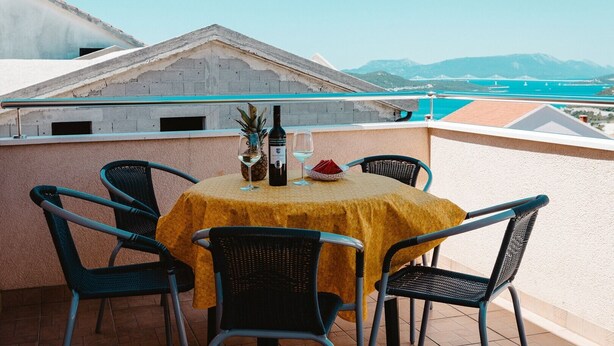 Une table recouverte d’une nappe jaune, quatre chaises, une bouteille de vin et un ananas, le tout sur un balcon offrant une vue sur la mer et les montagnes.