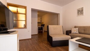 Un salon moderne avec un téléviseur à écran plat, un canapé, un fauteuil et une table basse. On aperçoit à l’arrière-plan un coin repas avec une table et des chaises.