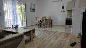 Ein modernes Wohnzimmer mit einer Couch, einem Esstisch mit Stühlen, einer Küchenzeile inklusive Kühlschrank und Backofen sowie einem großen Fenster mit Vorhängen.