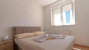Una cama bien hecha con cabecera color crema, una mesita de noche con lámpara y una ventana con persianas.
