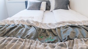 Ein Bett mit grau-weißer Decke, ein Fenster mit Blick auf Grünflächen und eine weiße Wand.