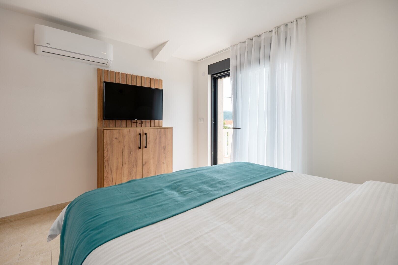 Une chambre d’hôtel avec un lit, un téléviseur fixé à un meuble en bois et une fenêtre ornée de rideaux légers.