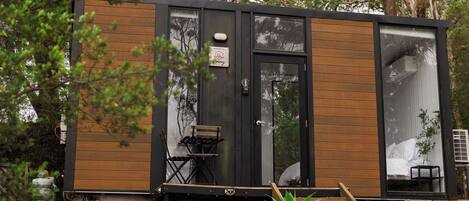 A modern, compact mobile home with a wooden exterior, a sliding door, and a small porch area with a bench and a table.