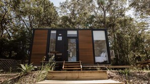 A modern, compact cabin with a dark exterior, large windows, and a wooden deck in front.
