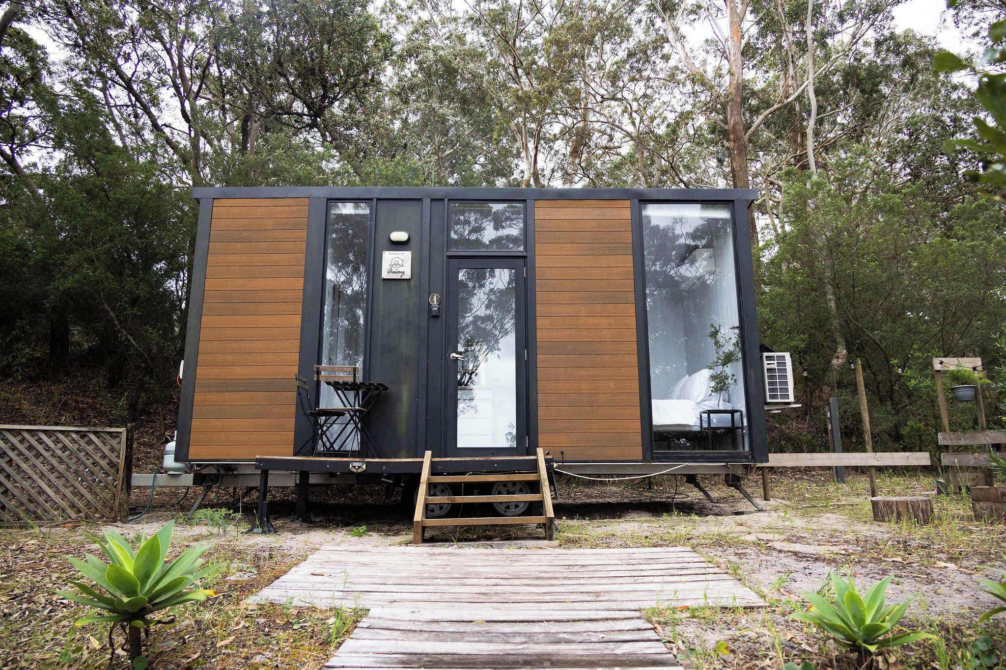 Una cabaña pequeña y moderna con fachada de madera y ventanas amplias, ubicada en zona boscosa y con una terraza de madera al frente.