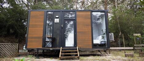 A small, modern cabin with a wooden exterior and large windows, situated in a wooded area with a wooden deck in front.
