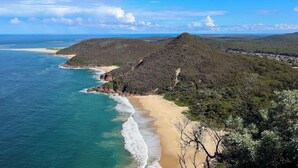 A coastal landscape with a beach, forested hills, and a clear sky.