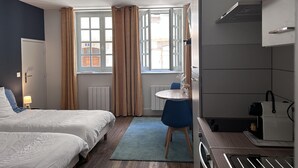 Ett hotellrum med två sängar, ett skrivbord, en stol, ett fönster med gardiner och ett litet kök.