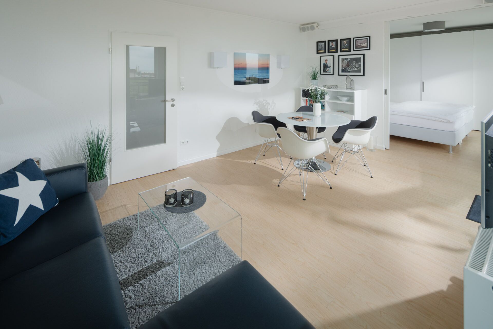 A modern living room with a glass dining table, black and white chairs, a grey rug, and a black sofa with a white star pillow.
