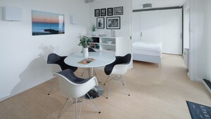 Ein moderner, hell erleuchteter Raum mit einem runden Esstisch, zwei Stühlen, einem weißen Sofa und einem Bett. An der Wand befinden sich gerahmte Bilder und auf dem Tisch steht ein Blumentopf.