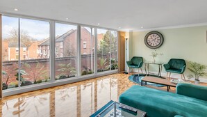 Ein geräumiges Wohnzimmer mit großen Fenstern, einem grünen Sofa, zwei Sesseln, einem kleinen Tisch und einer Wanduhr.
