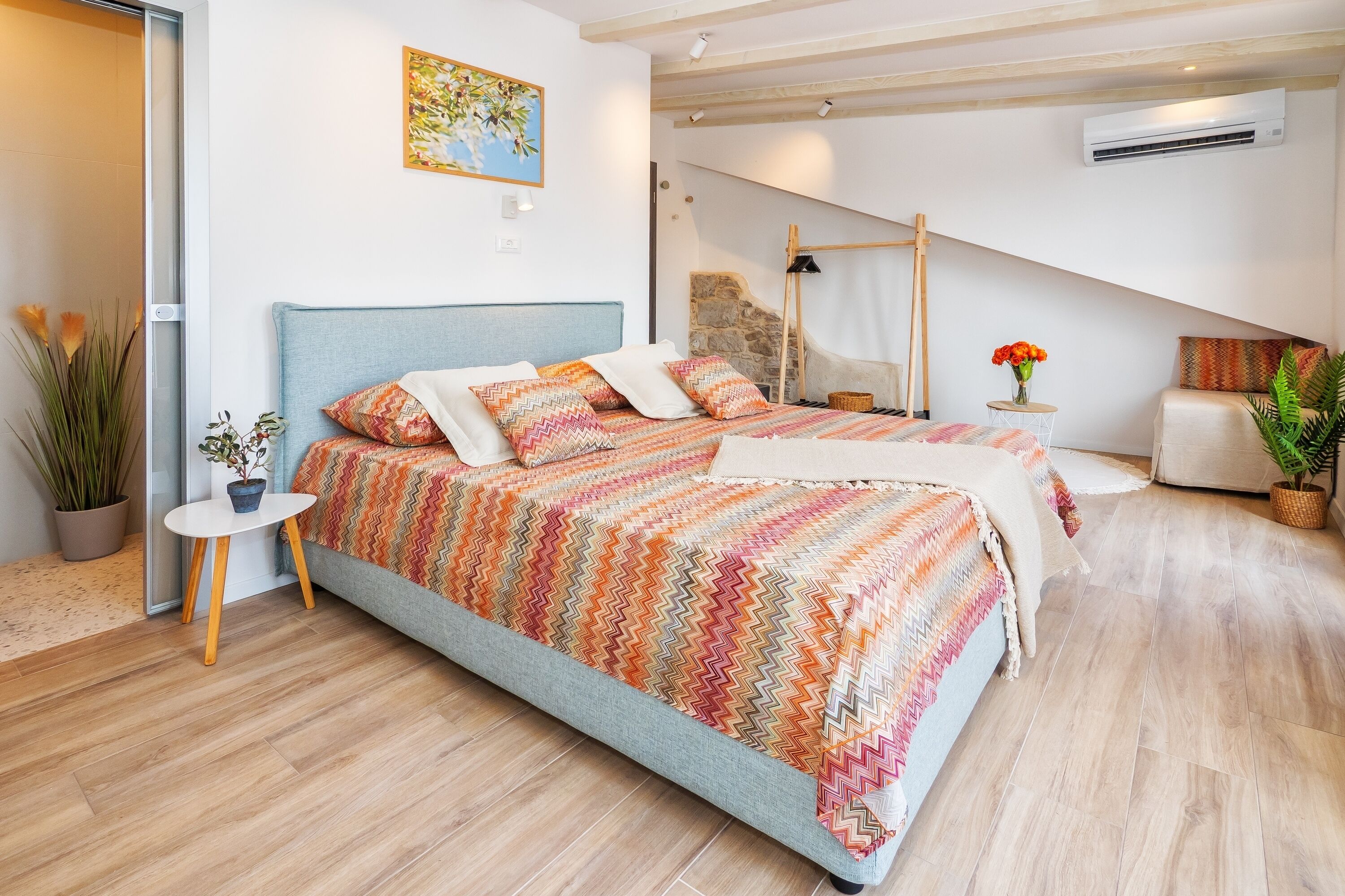 Une chambre à coucher moderne avec un grand lit, une petite table de chevet, une plante en pot et un tableau accroché au mur.