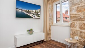 A modern room with a large TV displaying a coastal cityscape, a white console table with a plant, a stone wall, and a window with a view of buildings.