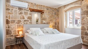 A bedroom with a stone wall, a bed with white bedding, a bedside table with a lamp, and a window with curtains.