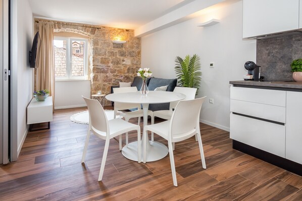 Une cuisine moderne avec des meubles blancs, une table de salle à manger ronde entourée de chaises blanches, un mur en pierre et une fenêtre avec des rideaux.