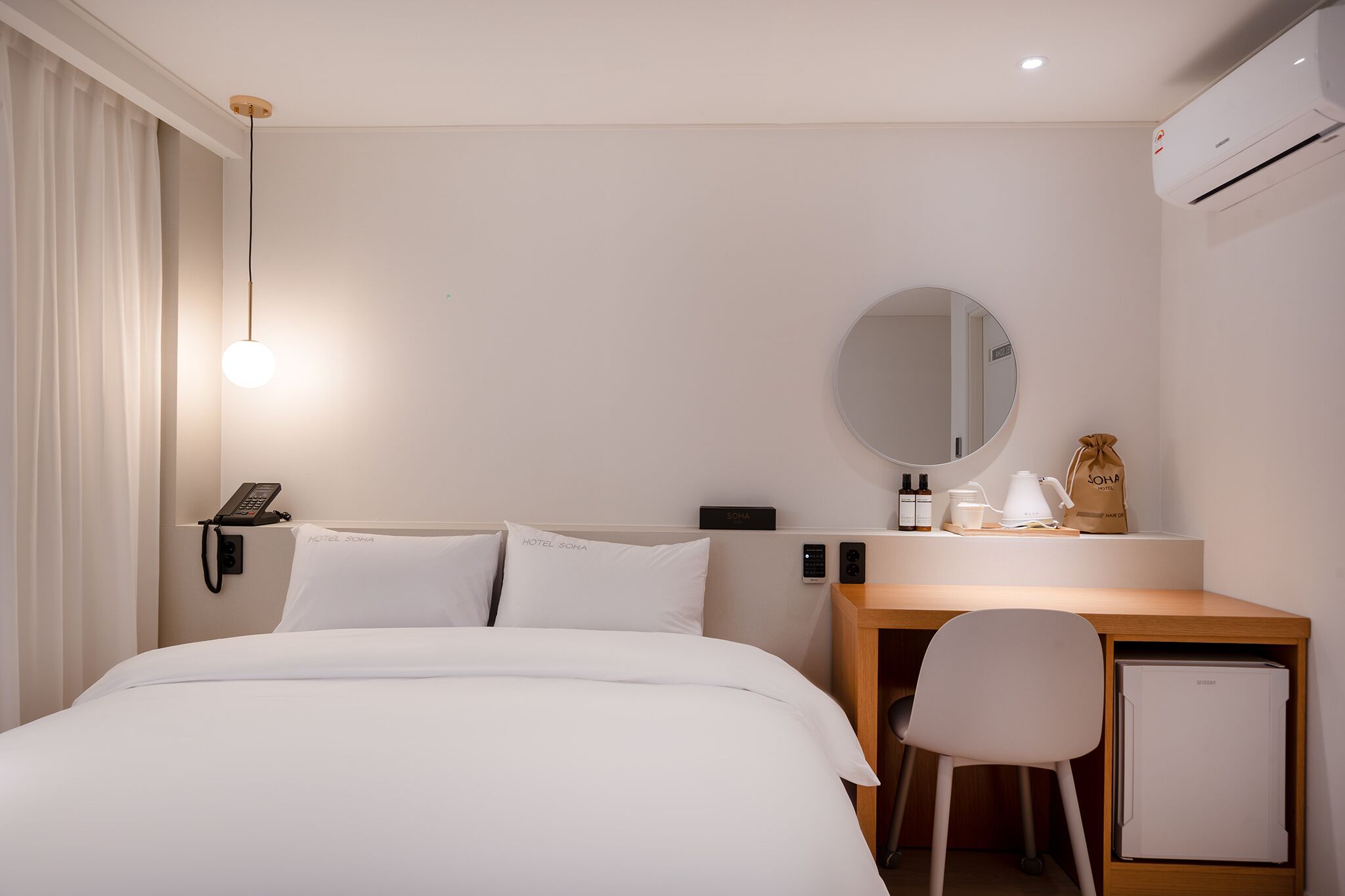A hotel room with a bed, a desk, a chair, a mirror, a phone, a coffee maker, a bag, and a mini fridge.