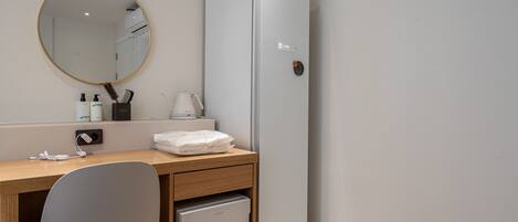 Habitación pequeña con escritorio de madera, un refrigerador compacto, espejo y lavamanos.