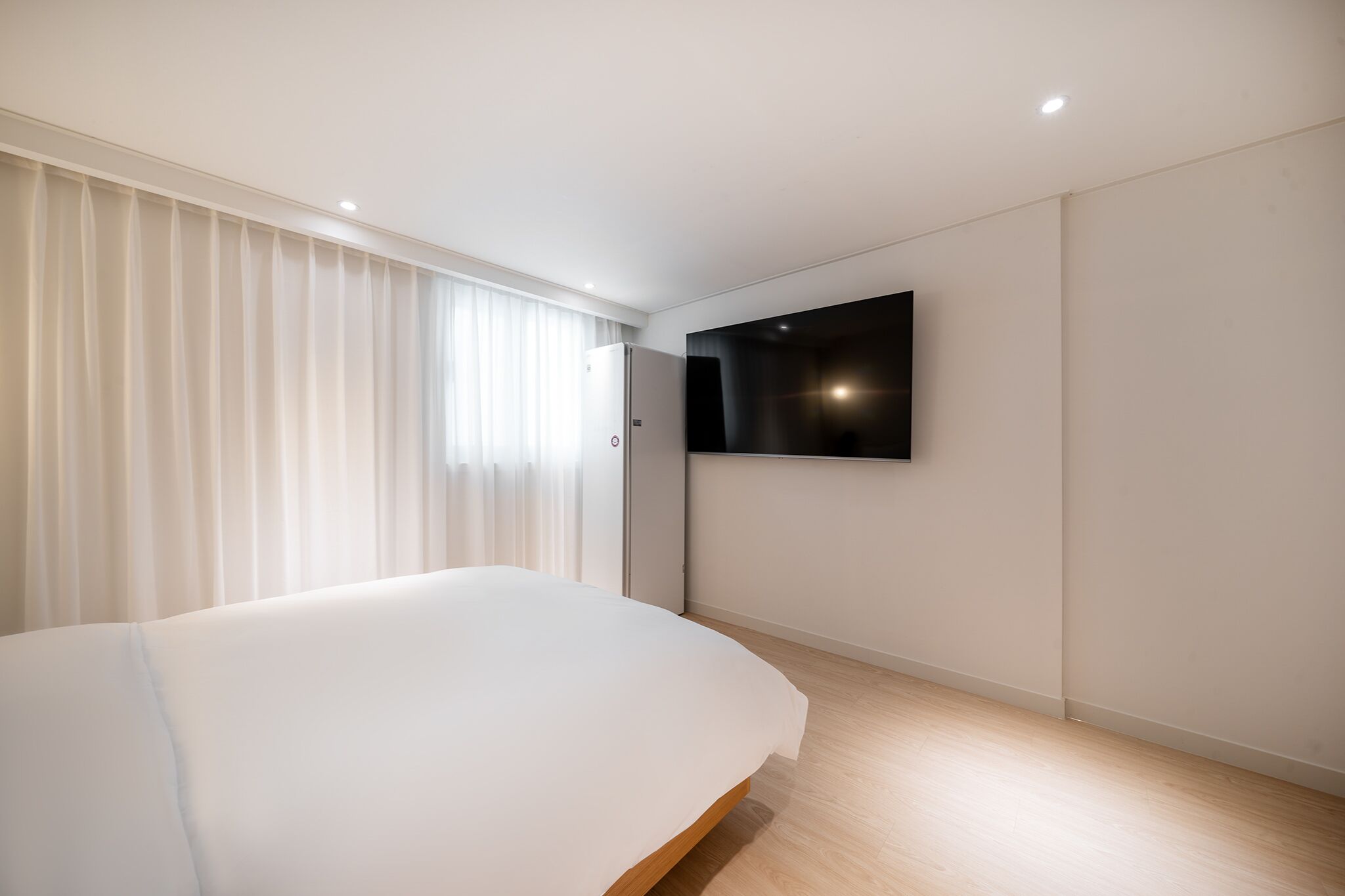 A modern bedroom with a large bed, a flat-screen TV mounted on the wall, and a window with sheer curtains.