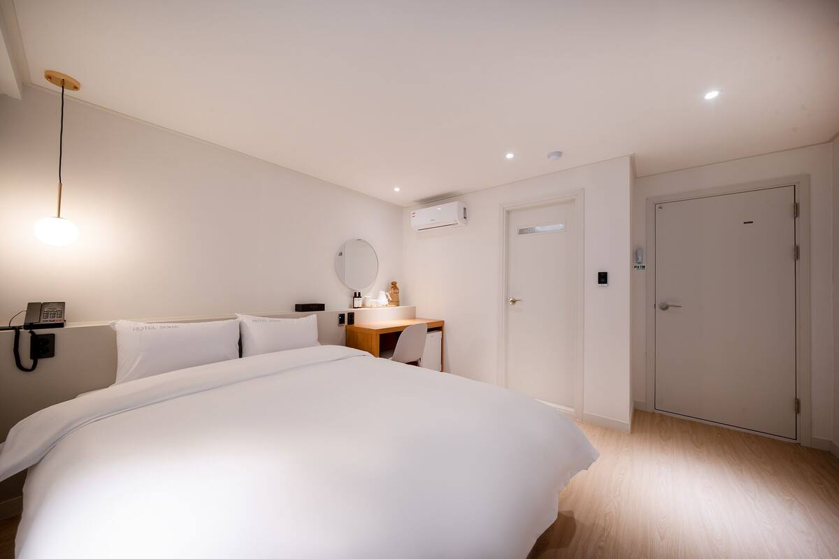 A hotel room with a large bed, a desk with a chair, a round mirror, an air conditioning unit, a phone, and a door.