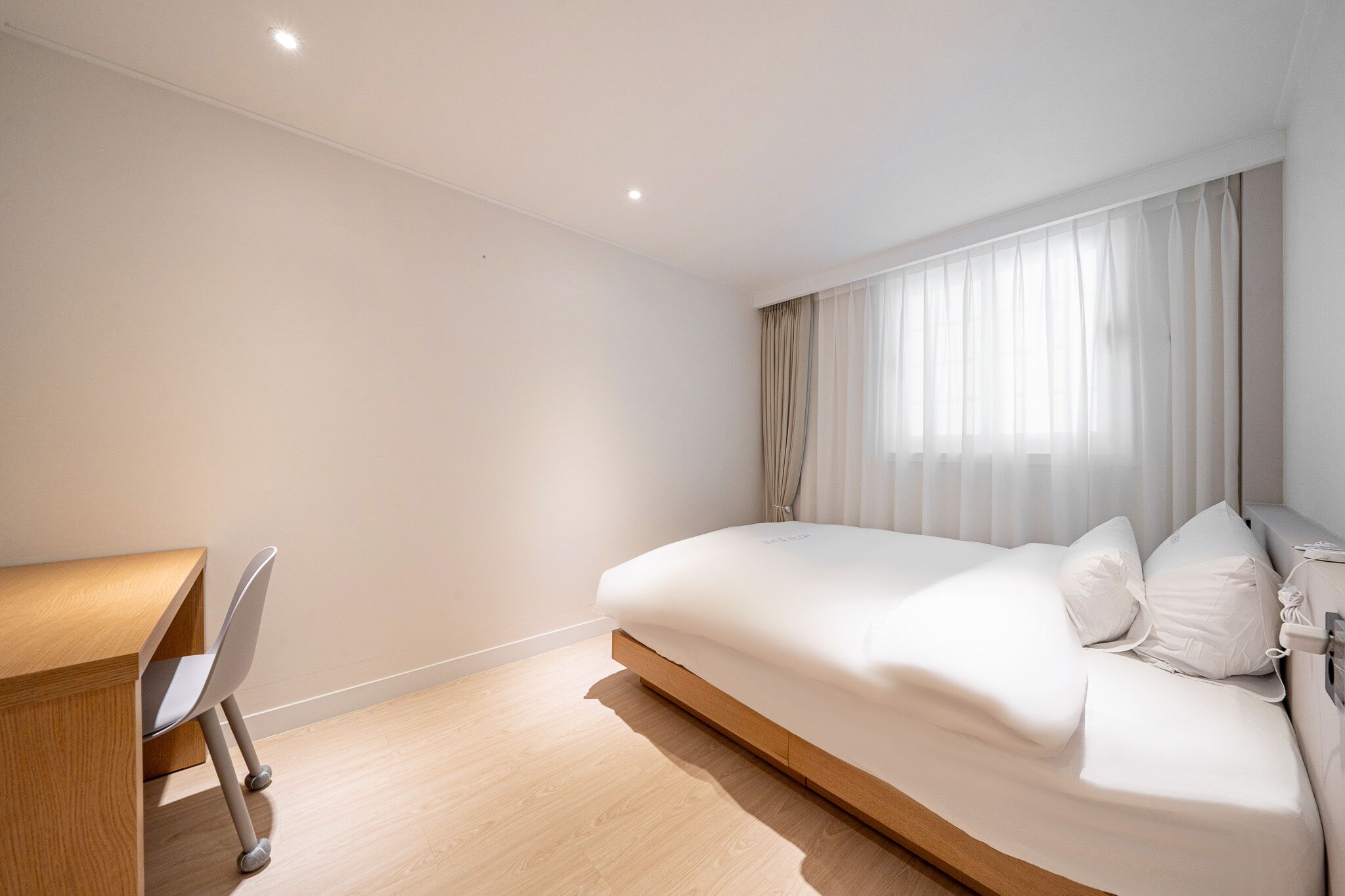 A modern hotel room with a wooden desk, a bed with white bedding, and sheer curtains on the window.