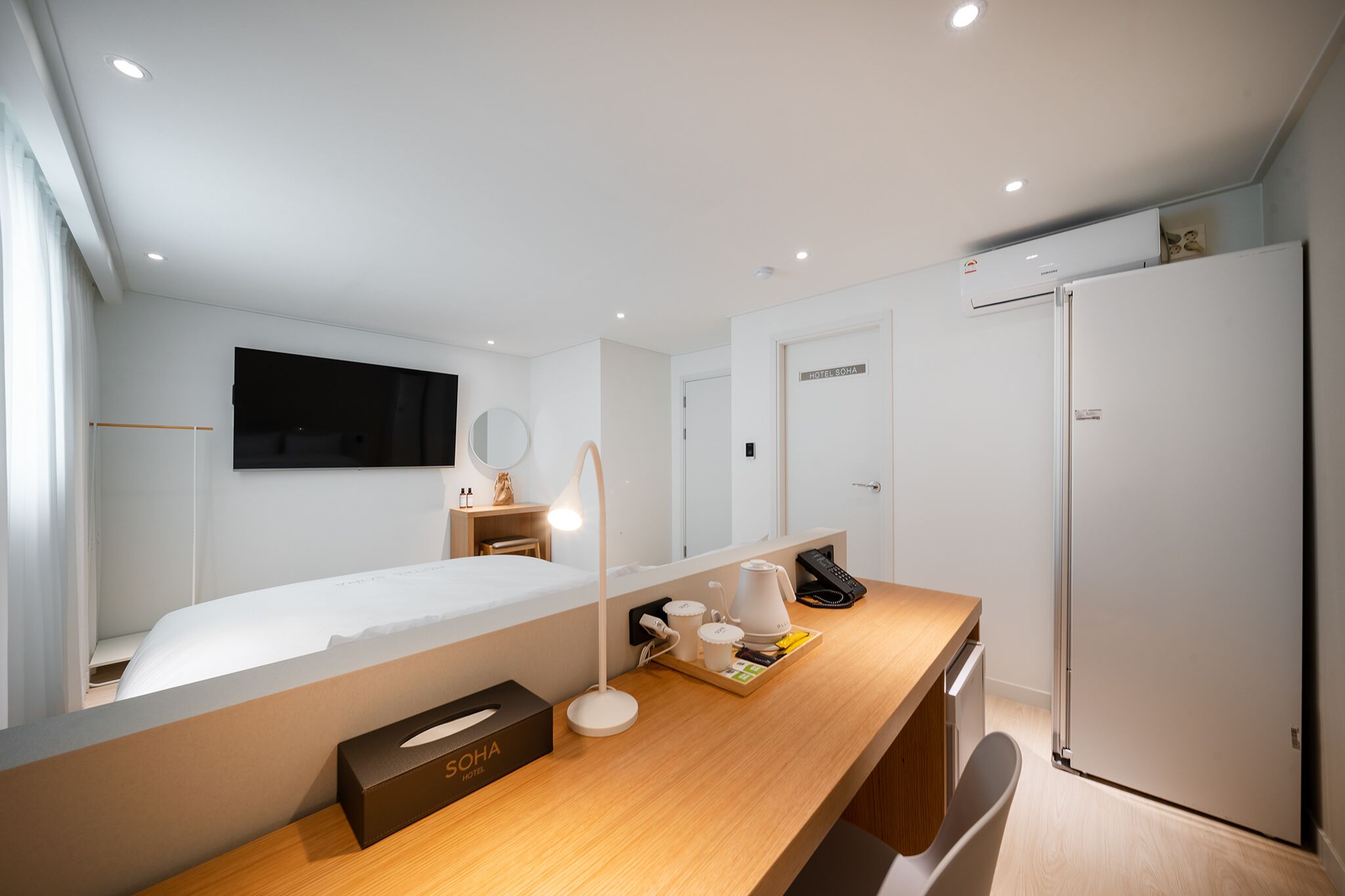 Una habitación de hotel moderna con cama, mesita de noche, escritorio con lámpara, espejo, televisor montado en la pared y refrigerador.