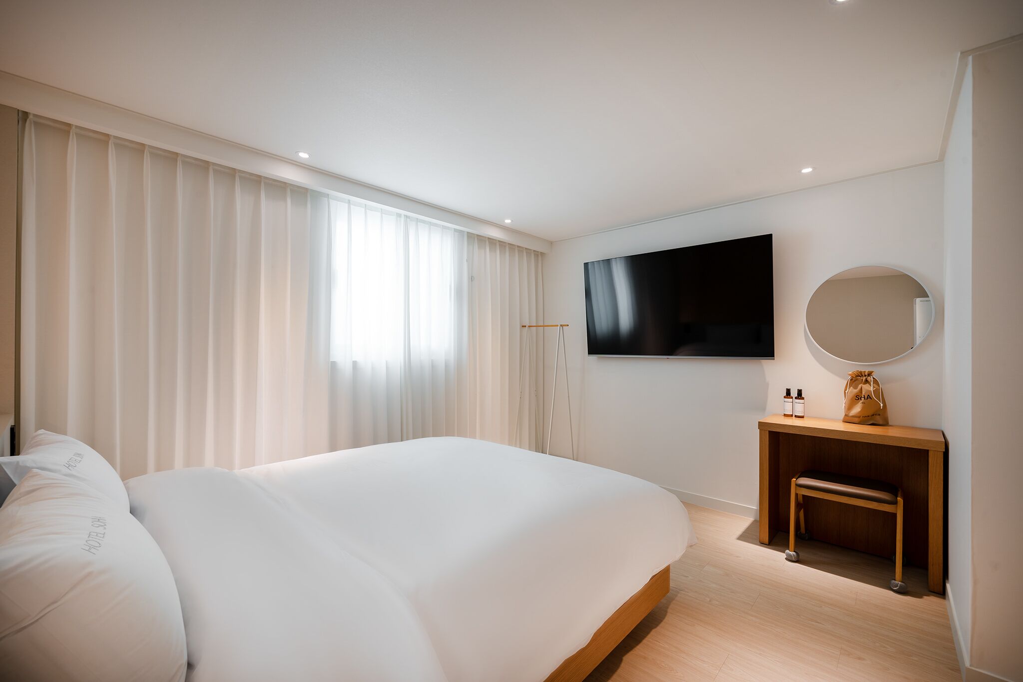A modern hotel room with a large bed, a TV mounted on the wall, a small wooden bench, and a bedside table with a mirror and two bottles.