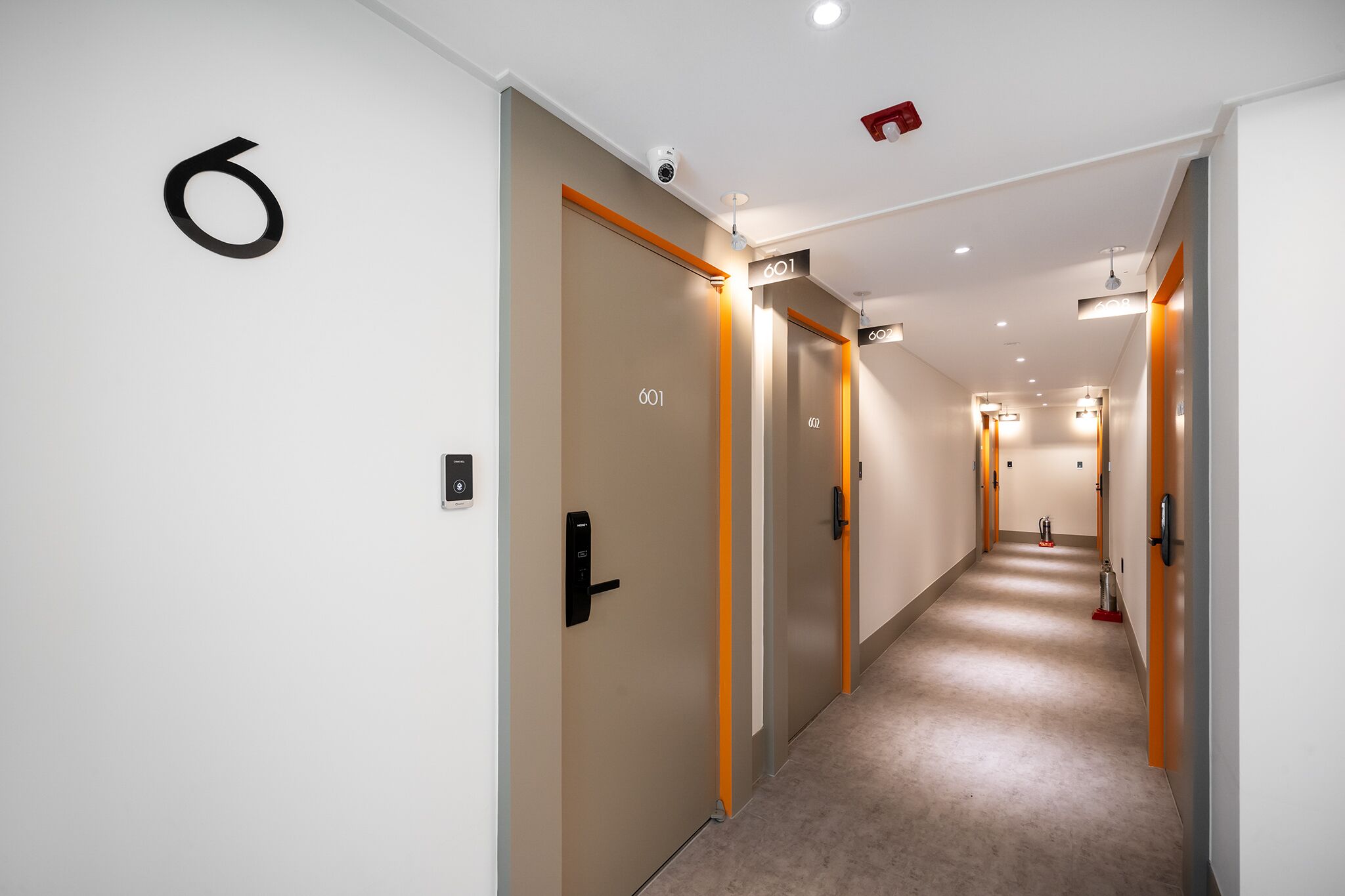 A modern hotel corridor with numbered doors, a security camera, and a fire extinguisher.